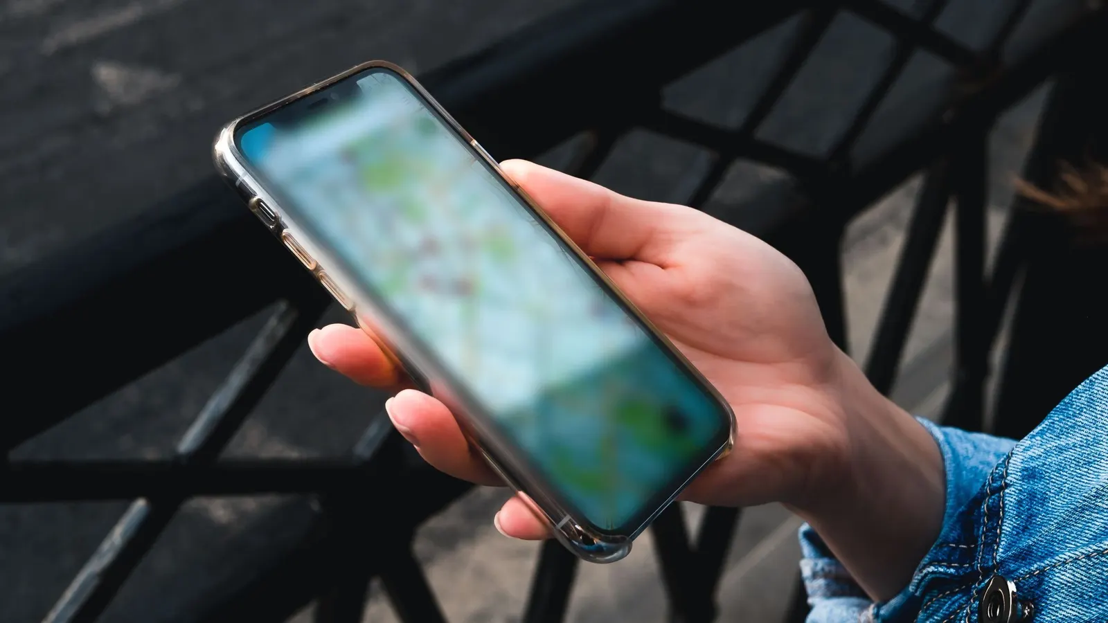 Eine Hand hält ein Smartphone mit Kartenansicht im Freien vor einem dunklen Geländer, daneben ist der Ärmel einer Jeansjacke zu sehen.