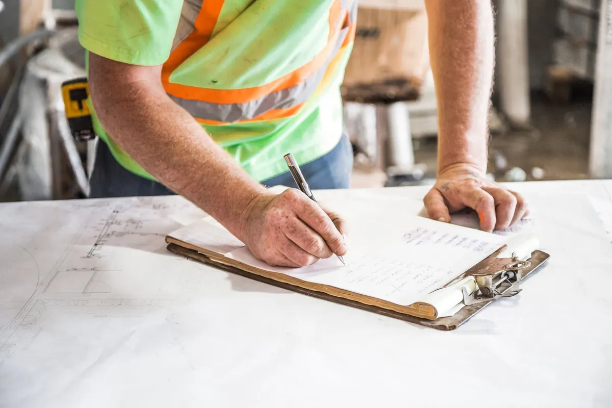 Ein Handwerker in Warnweste schreibt mit einem Stift auf ein Klemmbrett, das auf einem Bauplan auf der Baustelle liegt.